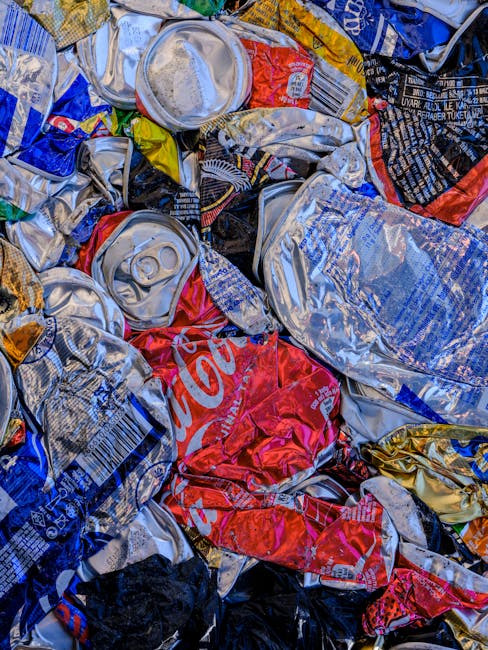 A close-up view of a large pile of crushed aluminium cans and plastic bottles, mixed with various colours and printed labels, stacked haphazardly on a flat surface. The cans, primarily silver with some featuring red, blue, and yellow branding, show dents and creases from being flattened. The plastic bottles are crumpled with some labels visible, including a prominent red Coca-Cola logo. The scene appears to be part of waste ready for collection or recycling, typical of waste management services such as those provided by Rubbish Collection Enfield. The background suggests an indoor or outdoor environment associated with waste handling, with no clear landscape or surroundings visible. The lighting highlights the metallic and plastic surfaces, emphasizing the textures of compressed materials prepared for disposal or recycling, in a manner consistent with private waste collection rather than municipal systems. This scene illustrates the accumulation of recyclable waste and highlights the importance of efficient rubbish removal services in the Enfield area.