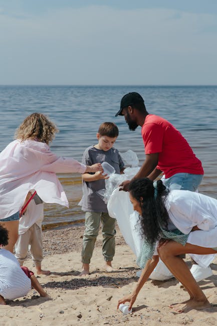 A diverse group of people participating in a beach cleanup, with a woman in a pink jacket and white pants handing a plastic bag to a young boy in a grey t-shirt, while an adult man in a red t-shirt and cap helps collect rubbish. A woman with curly black hair in a white shirt is bending down to pick up debris, and a child in a striped shirt crouches nearby on the sandy shore. The background features calm sea waters under an overcast sky, with the shoreline scattered with captured waste and discarded plastic items. The scene depicts communal efforts of waste collection and environmental conservation, illustrating an outdoor activity aligned with private rubbish removal services, such as those offered by Rubbish Collection Enfield, emphasizing community involvement in waste management.