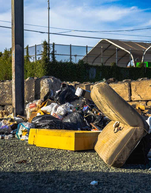 Two large black plastic rubbish bags and a rectangular cardboard box are placed on a curbside pavement in front of a dark metal fence with vertical bars and a brick wall behind it. The bags appear filled with waste material, showing crumpled and uneven surfaces, with some reflective surfaces indicating plastic material. The cardboard box, partially torn, has printed text on its surface and is positioned leaning against one of the bags. The pavement is adjacent to a road, with a curb separating them, and a hint of greenery from a tree or bush is visible at the top left corner of the image. The scene suggests an area awaiting collection or disposal, with natural lighting casting soft shadows, and the overall tone is neutral and unembellished, consistent with private rubbish collection services or alternative waste handling scenarios in an urban environment, possibly managed by an independent waste removal provider such as Rubbish Collection Enfield.