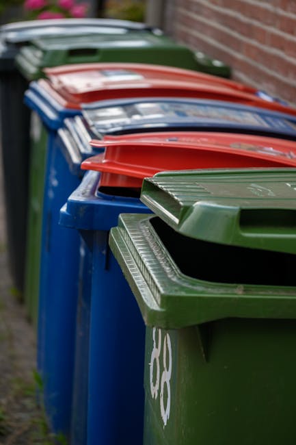 A row of large wheelie bins for rubbish collection, positioned along a pavement beside a brick wall. The bins are made of rigid plastic with textured surfaces and are different colours, including blue, green, and red, indicating various waste categories. The green bin in the foreground has a slightly open lid, revealing the interior, while the other bins have lids closed and are stacked or leaning slightly. The bins are placed on a concrete ground area, with a narrow strip of grass visible on the left, and the background shows a blurred view of a brick wall and a vehicle parked further down the pavement. The scene represents a typical setting for external waste collection or private rubbish removal services, with the branding of Rubbish Collection Enfield occasionally present on the bins or visible nearby.