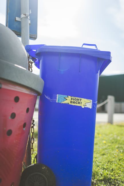 A tall, bright blue plastic rubbish bin with a closed lid and a handle on top, positioned outdoors on a grassy area, with a small yellow and blue sticker that reads 'Fight Back!' affixed to its front. To the left of the blue bin, part of a red container with black circular holes and a grey lid is visible, along with a white pole and chains attached to the red container. The background includes a blurred view of a road, some fencing, and a partly cloudy sky, suggesting the scene is at a roadside or service area. The image captures the typical outdoor setting for private waste collection, emphasizing the use of independent rubbish containers for waste disposal services like those offered by Rubbish Collection Enfield, aligning with the theme of alternative waste handling and rubbish removal solutions.