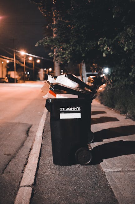 A black waste bin with a white label that reads 'St. John's' is positioned on the side of a quiet street at night. The bin is filled with assorted rubbish, including paper and cardboard, which is overflowing and spilling onto the surrounding area. The street is illuminated by overhead streetlights, casting warm light on the scene, with a slight glow reflecting off the curb and pavement. Behind the bin, there are trees and bushes along a hedge line, and faintly visible parked cars further along the road. The surface of the street and sidewalk is made of asphalt and concrete, respectively, with shadows cast by the bin and nearby foliage. The scene appears to depict an outdoor rubbish collection spot, possibly part of a private waste disposal arrangement managed by Rubbish Collection Enfield, illustrating typical on-site waste accumulation in residential or commercial areas.