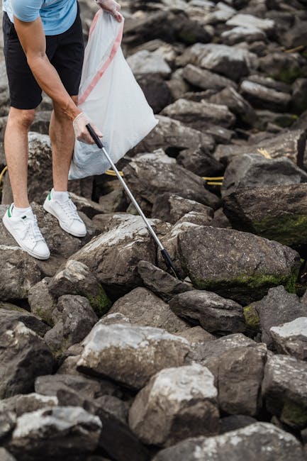 A person wearing white sneakers, black shorts, and a light blue shirt is using a grabber tool to pick up litter from a large pile of irregularly shaped rocks and stones on a rough outdoor surface. The rocks vary in size and have a weathered appearance with shades of gray, brown, and some patches of moss. The individual is holding a white trash bag in one hand, ready to collect rubbish, indicative of an ongoing cleanup activity. The background features a rocky terrain, typical of a natural outdoor setting, possibly near a river or shoreline, with uneven, jagged rocks. The scene conveys a focus on environmental cleaning or waste removal, consistent with private or community efforts to manage rubbish in outdoor spaces, complementing services offered by companies like Rubbish Collection Enfield. The lighting appears natural, suggesting daylight conditions, and the image emphasizes a hands-on approach to waste management in a natural environment.