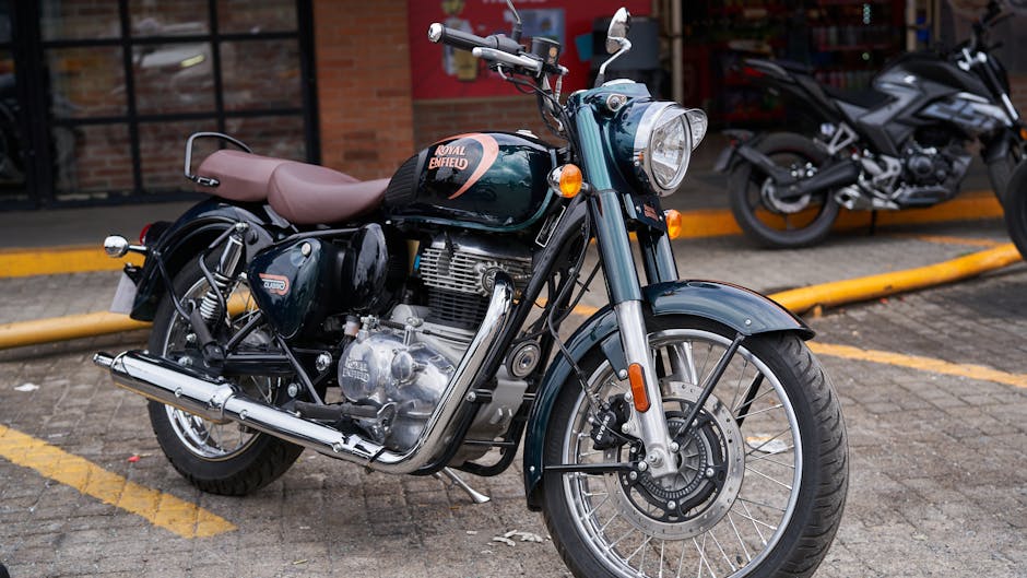 A close-up view of a dark green Royal Enfield motorcycle parked on a paved outdoor surface with yellow parking lines and a yellow curb. The motorcycle features a light brown leather seat, chrome exhaust pipe, and polished metal engine components. Its front wheel, with visible disc brake caliper, is turned slightly to the right, and the front fender matches the bike's body color. In the background, there are additional motorcycles, including a silver sports bike, parked adjacent to a brick building with large windows, suggesting an urban setting. The lighting appears natural, with soft shadows indicating daytime. This scene relates to private or independent vehicle storage in an area possibly used for collection or disposal purposes, aligning with the context of rubbish removal services such as those provided by Rubbish Collection Enfield for alternative waste management.