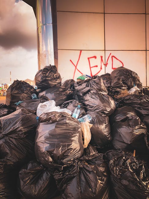 A large heap of black rubbish bags stacked against a modern building with a tiled exterior, with some filled with household waste and others appearing slightly torn or bunched. Among the bags, several clear plastic bottles and smaller pieces of debris are visible, adding to the cluttered appearance. In the background, part of the urban environment can be seen, with a partly cloudy sky overhead, suggesting natural daylight. Graffiti in red spray paint reading 'XEND' is visible on the wall behind the rubbish, indicating possible unauthorized waste disposal or a local environment issue. The scene captures an instance of waste accumulation that could be addressed through professional rubbish removal services, such as those provided by Rubbish Collection Enfield, highlighting the importance of proper waste management and adherence to local rubbish rules, especially in an urban setting where on-site clearance or alternative disposal methods may be necessary to maintain cleanliness and compliance.
